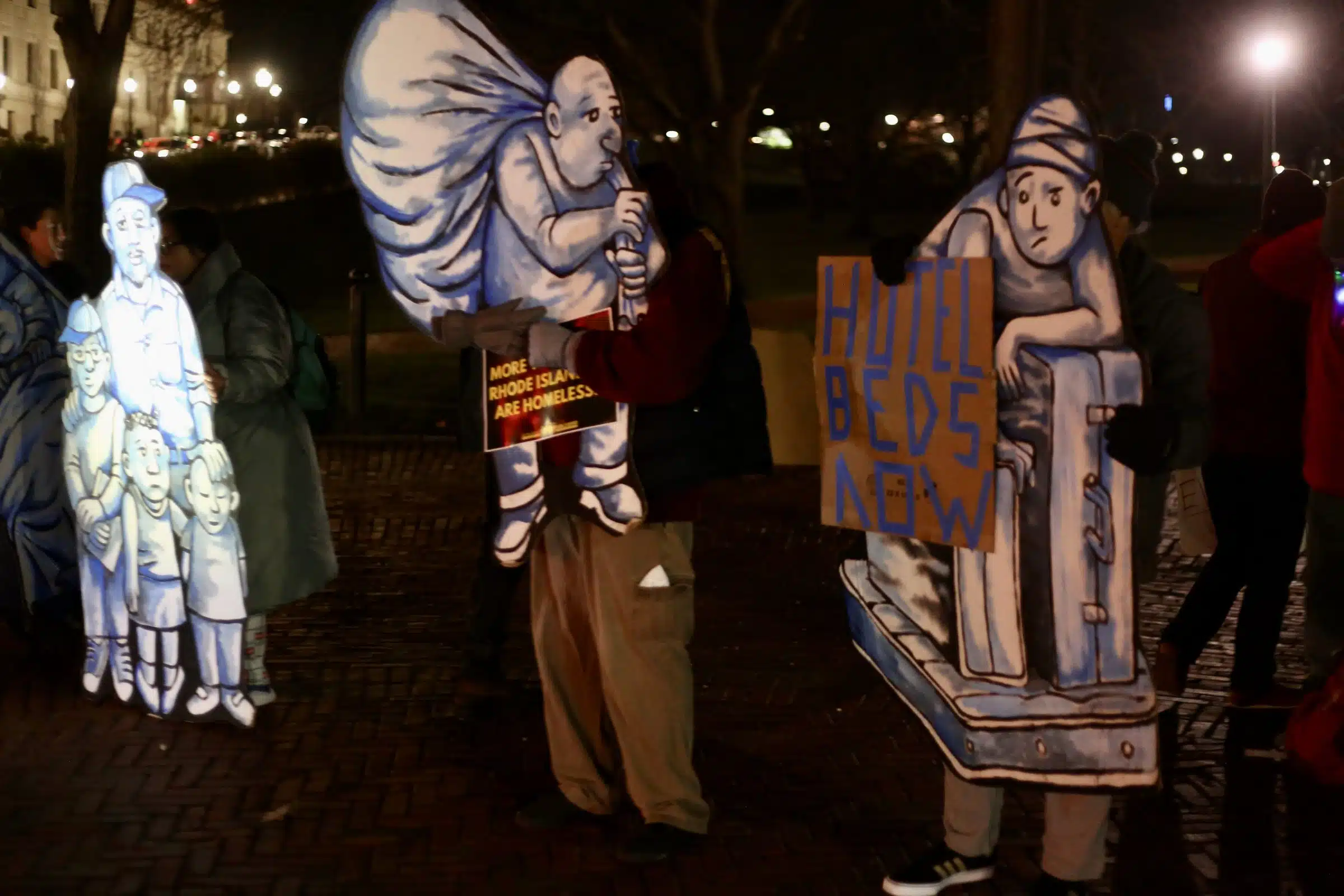 Life size cardboard cutouts of unhoused people in various states of homelessness on the lawn of the Rhode Island State House at night.