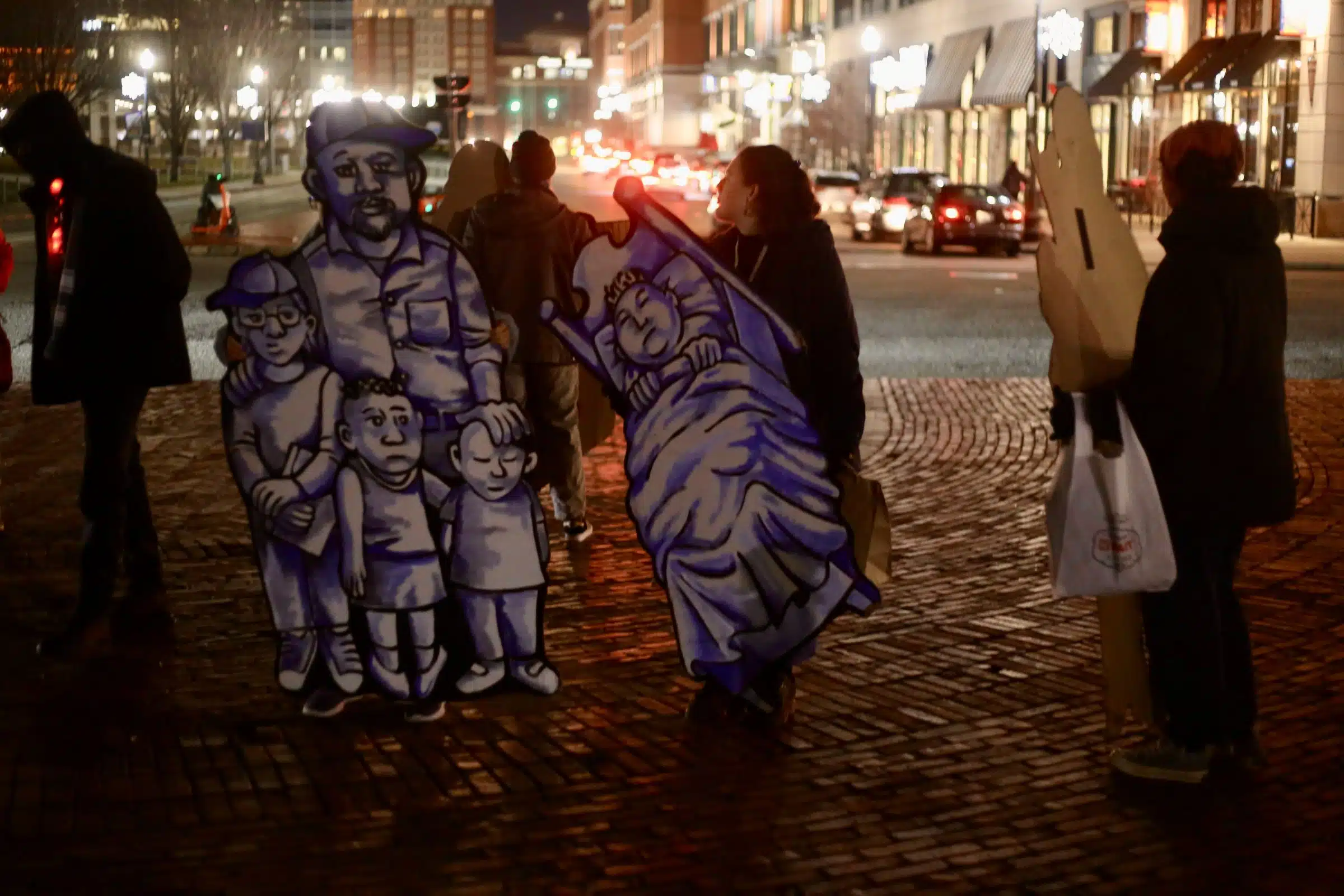 Life size cardboard cutouts of unhoused people in various states of homelessness on the lawn of the Rhode Island State House at night.