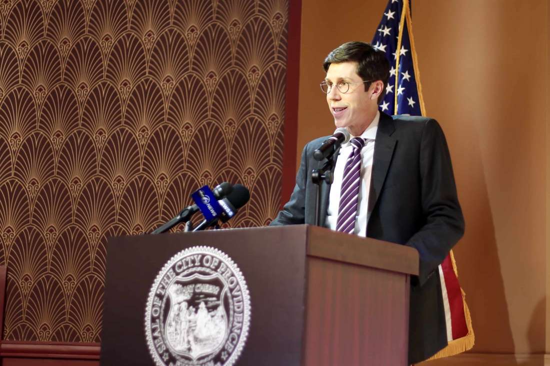 Providence Mayor Brett Smiley at a podium at the Providence Public Library, in front of an American flag and behind the Seal of the City of Providence.